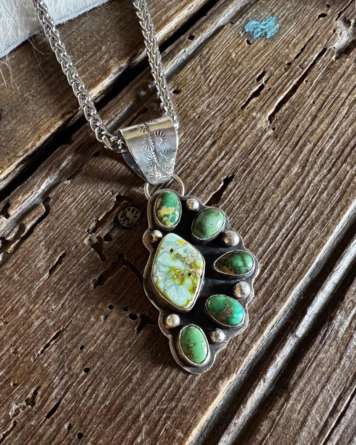 A silver-toned paw-shaped pendant necklace with green and white marbled turquoise-like stones, displayed on a rustic wooden surface with a white faux-fur prop.