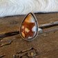 A teardrop-shaped ring featuring a brown and cream marbled cabochon stone in a silver-toned bezel setting, resting on a rustic wooden surface with a white fur trim in the background.