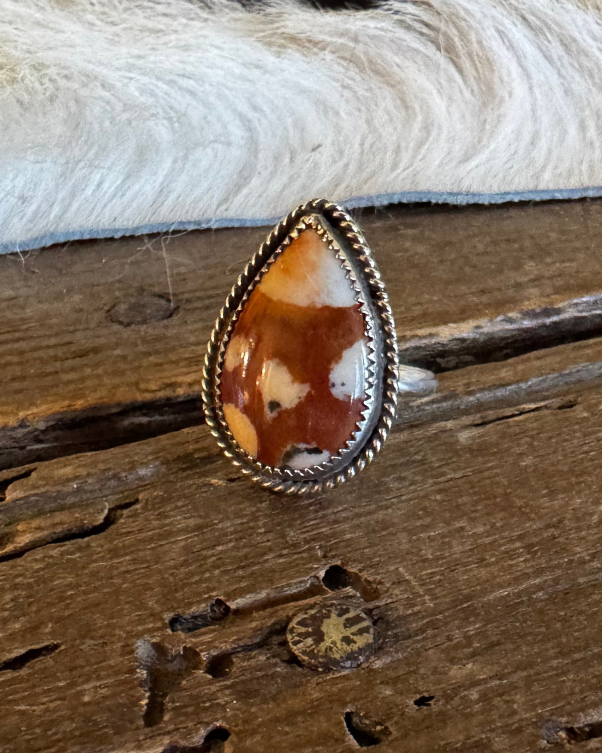 A teardrop-shaped ring featuring a brown and cream marbled cabochon stone in a silver-toned bezel setting, resting on a rustic wooden surface with a white fur trim in the background.