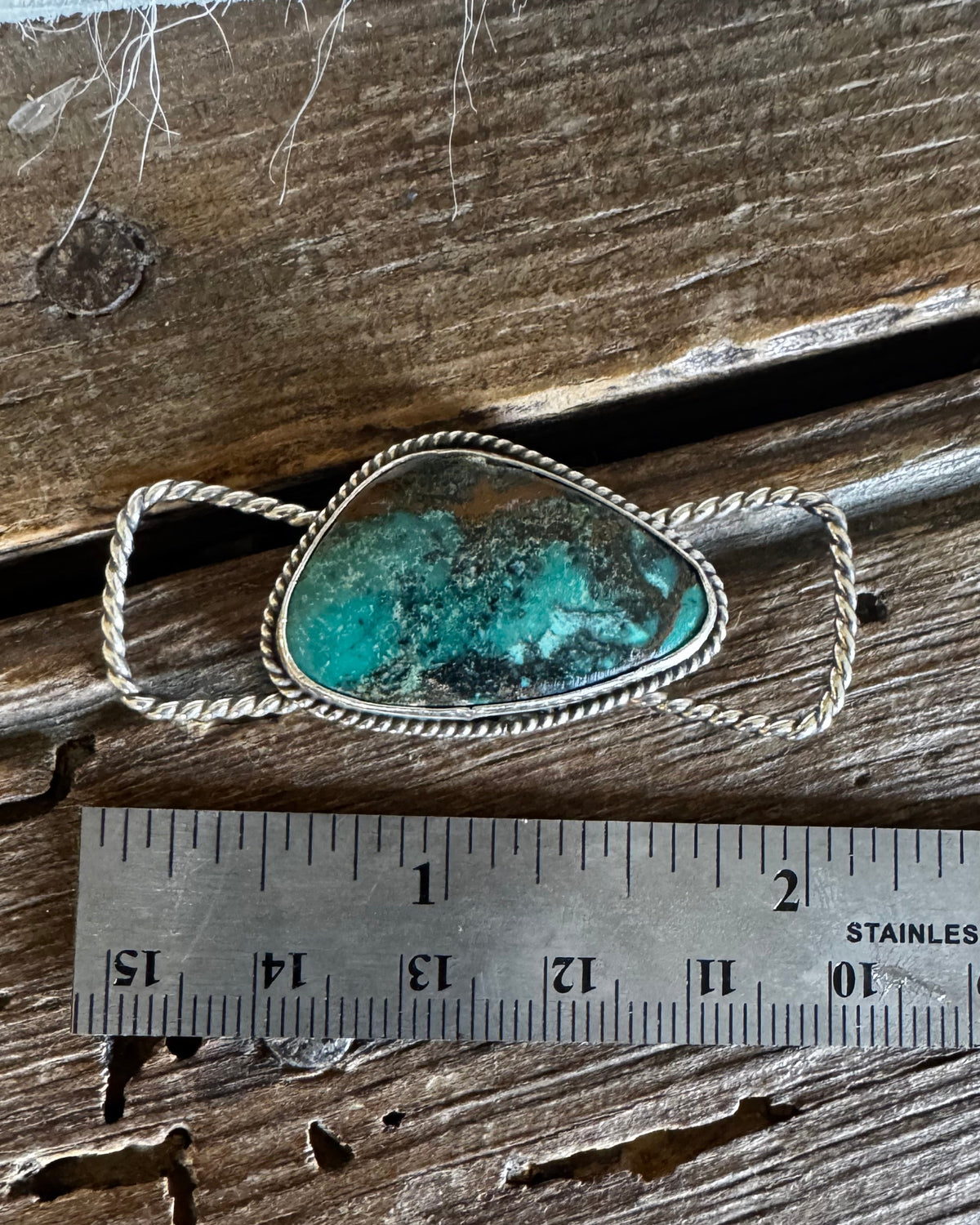 A silver-toned ring with a rough turquoise stone, shown on a rustic wooden surface next to a stainless steel ruler for scale.