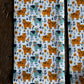 A close-up of a fabric product, likely a scarf or narrow textile panel, featuring a repeating pattern of orange and teal cows with yellow flowers and green leaves on a white background, styled next to a rustic dark wood surface.
