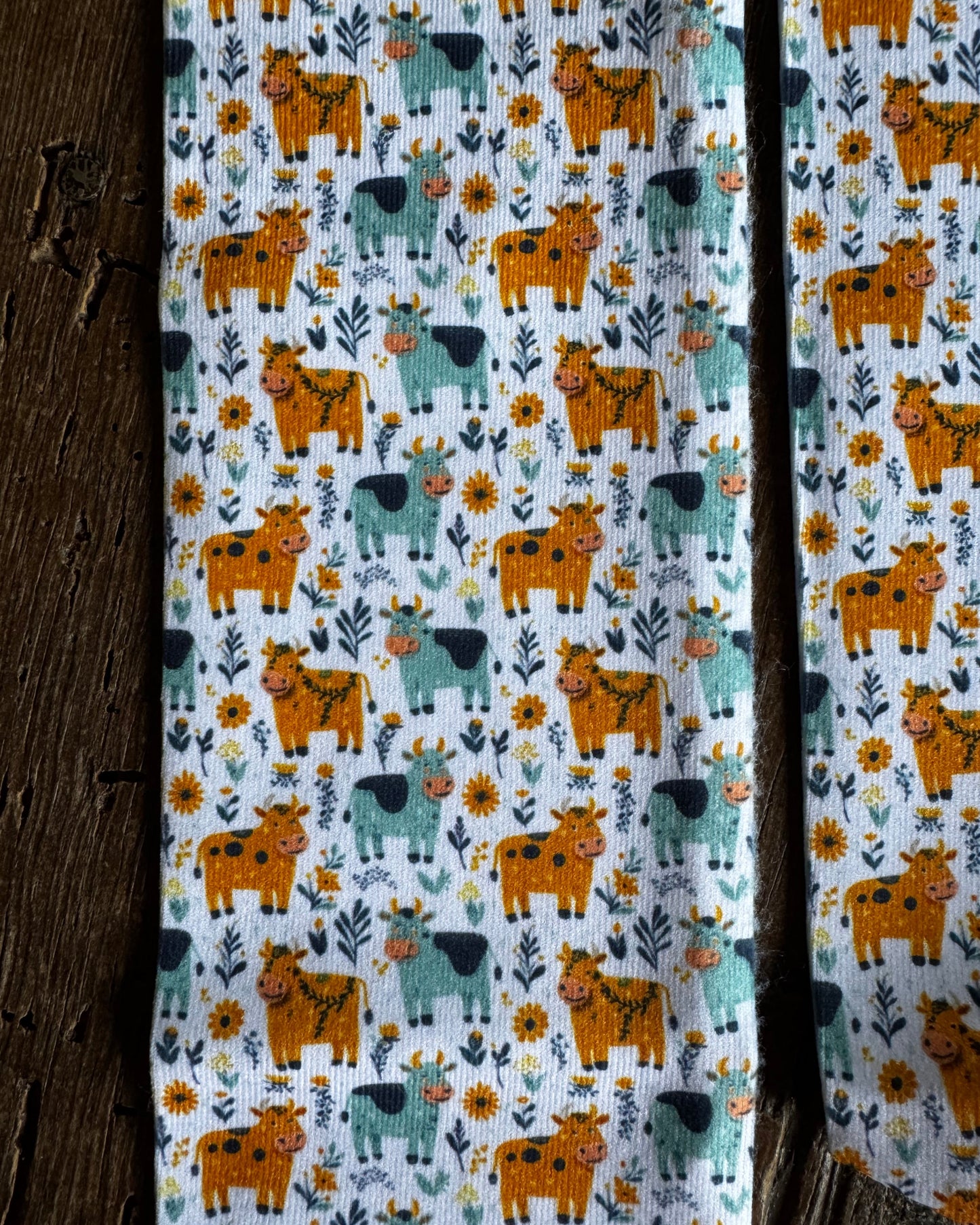 A close-up of a fabric product, likely a scarf or narrow textile panel, featuring a repeating pattern of orange and teal cows with yellow flowers and green leaves on a white background, styled next to a rustic dark wood surface.