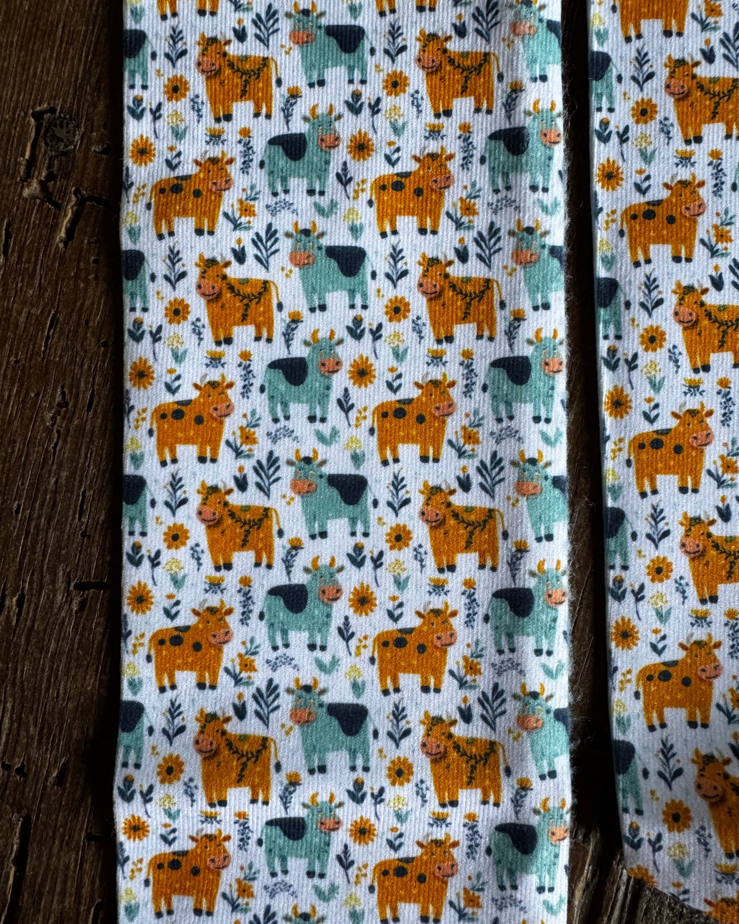 A close-up of a fabric product, likely a scarf or narrow textile panel, featuring a repeating pattern of orange and teal cows with yellow flowers and green leaves on a white background, styled next to a rustic dark wood surface.