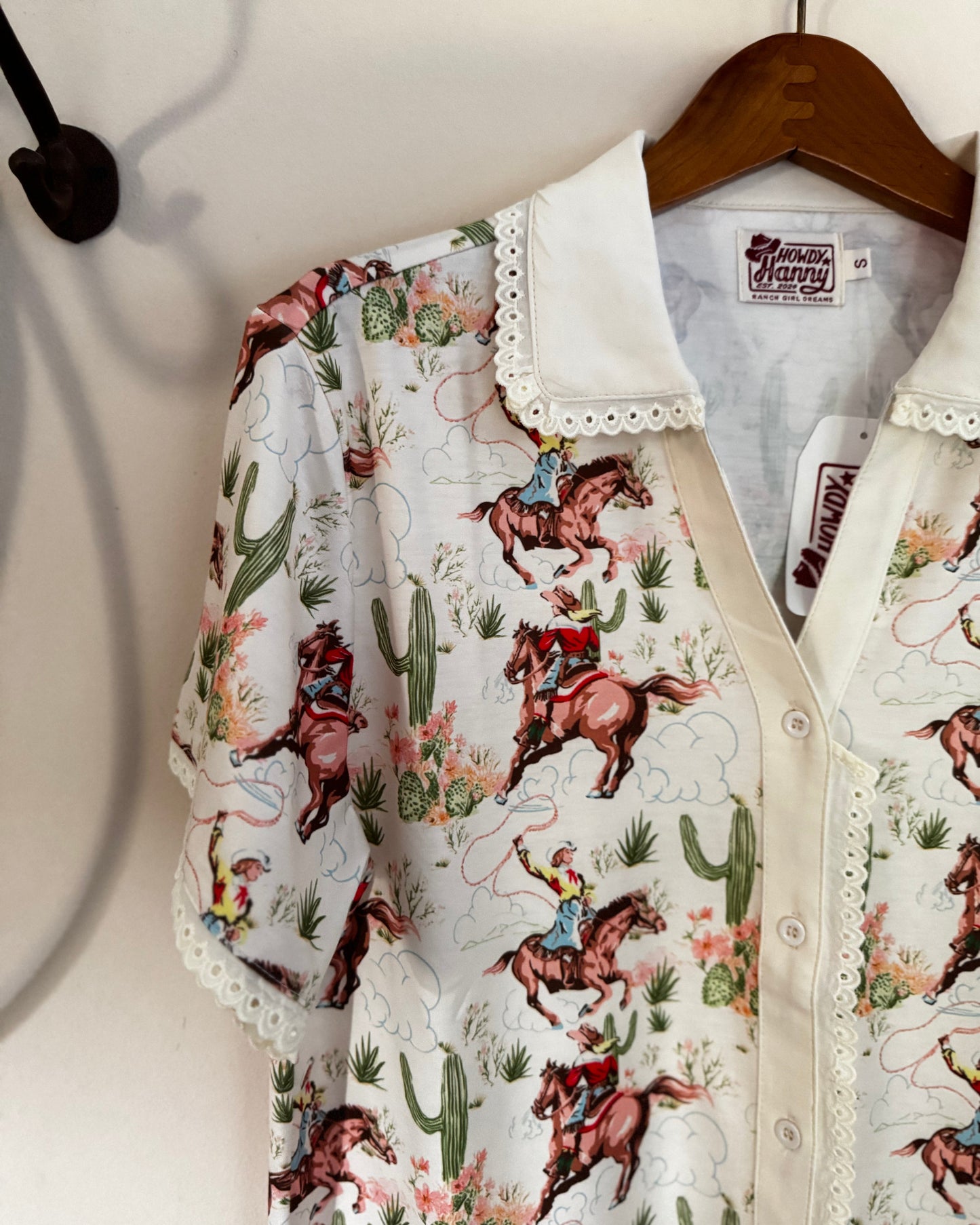 A short-sleeve button-up shirt with a white lace-trimmed collar, featuring a colorful western print of cowboy riders on horses with cacti and desert flowers. The shirt is displayed on a wooden hanger against a white wall.