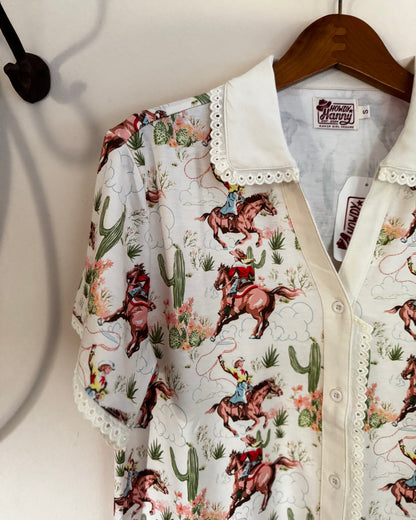A short-sleeve button-up shirt with a white lace-trimmed collar, featuring a colorful western print of cowboy riders on horses with cacti and desert flowers. The shirt is displayed on a wooden hanger against a white wall.