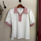 White blouse with red trim on a hanger against a beige wall.