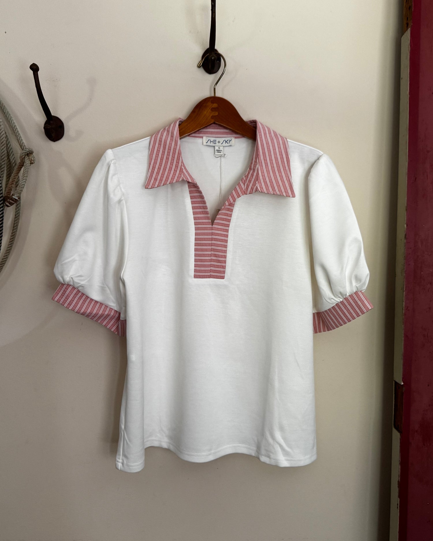 White blouse with red trim on a hanger against a beige wall.