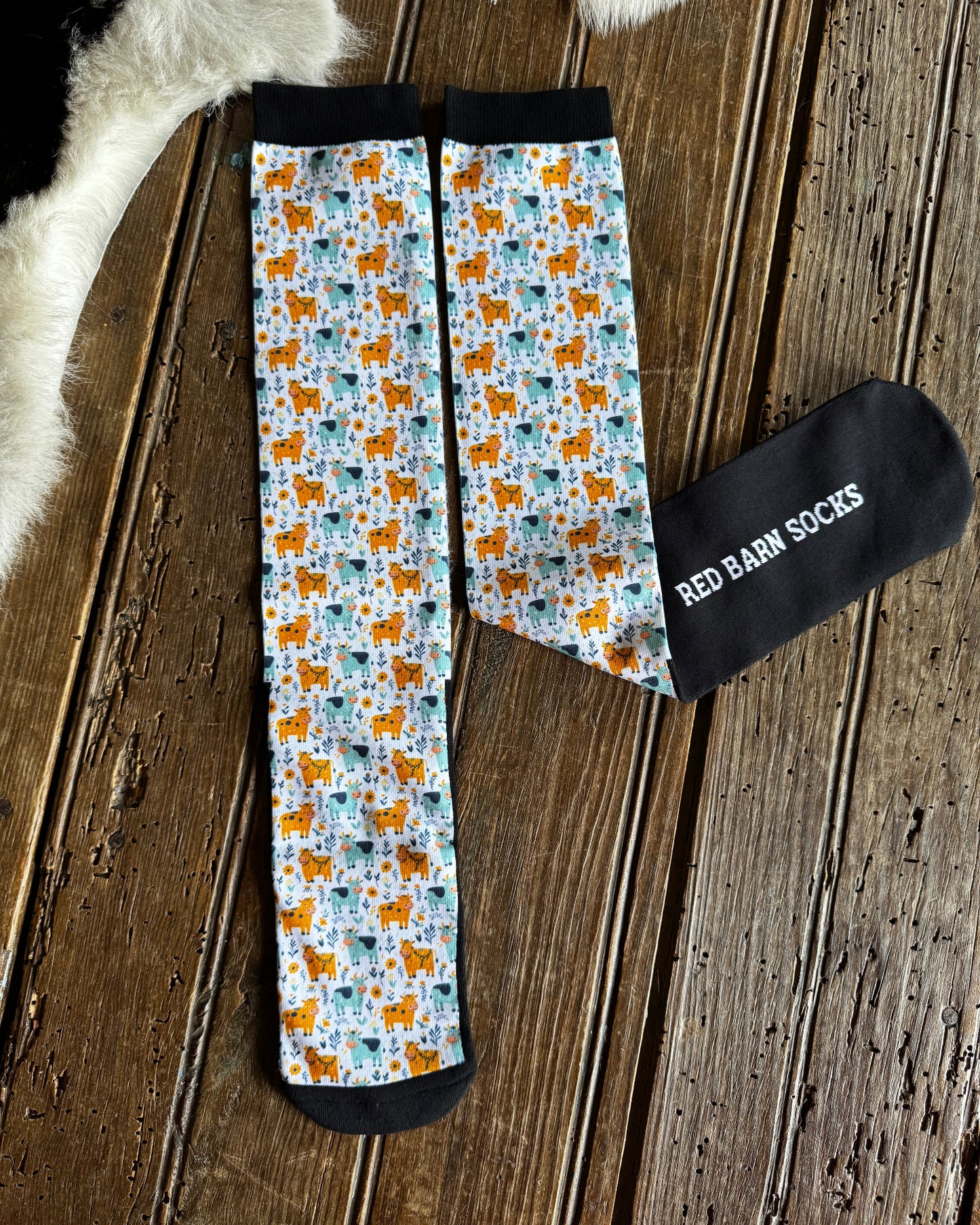 A pair of crew socks with a light blue background and an all-over pattern of orange dogs and teal birds, laid flat on a rustic wooden surface next to a black sock tag that reads 'RED BARN SOCKS'.
