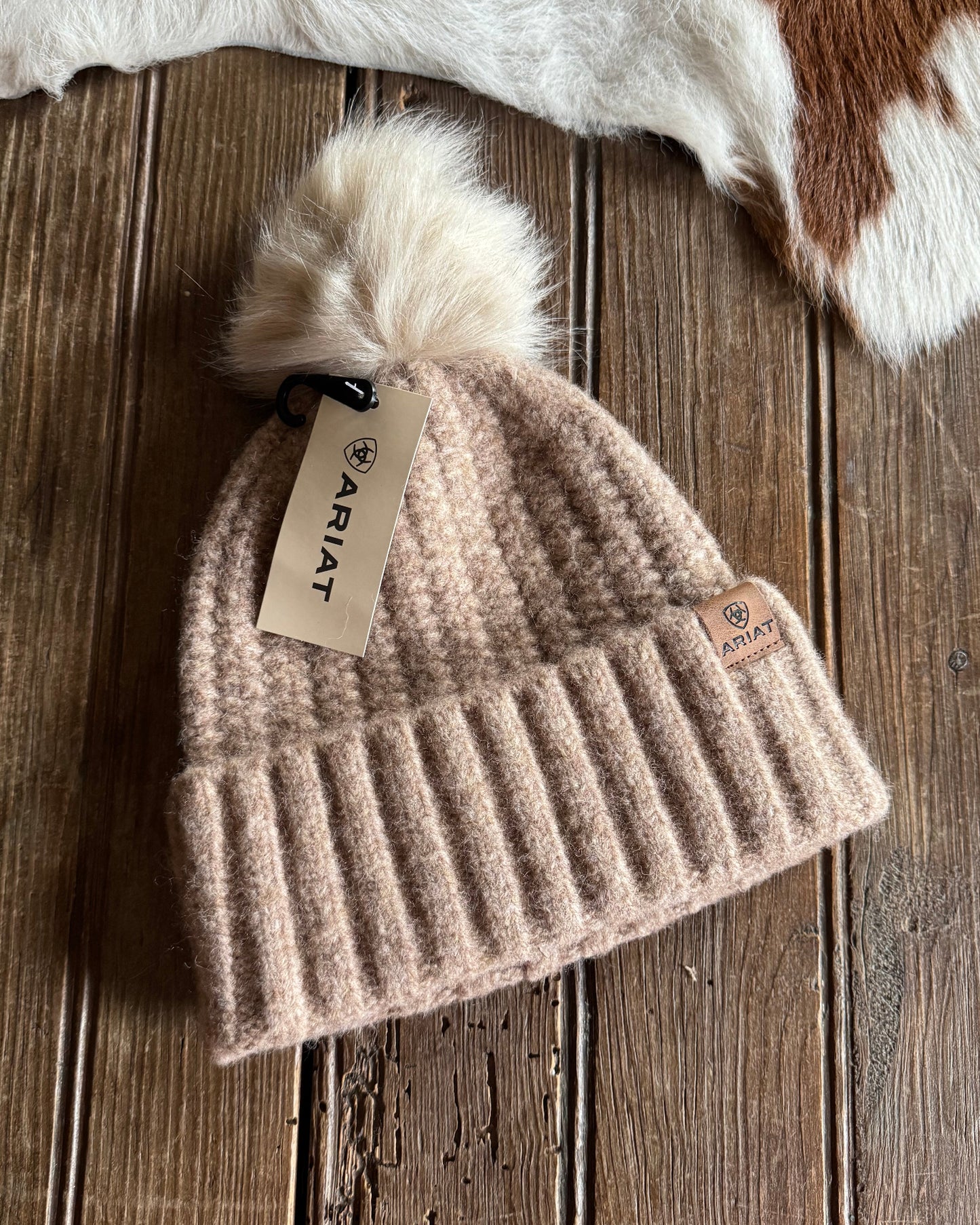 Beige knit beanie with pom-pom and Ariat tag on a wooden surface