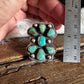 A hand holds a silver ring with turquoise stones next to a metal ruler over a rustic wooden surface.