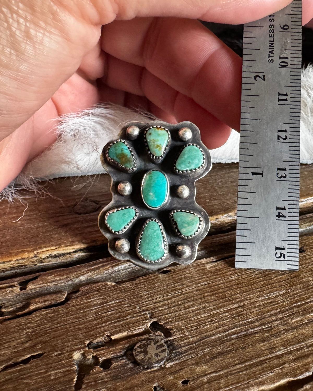 A hand holds a silver ring with turquoise stones next to a metal ruler over a rustic wooden surface.