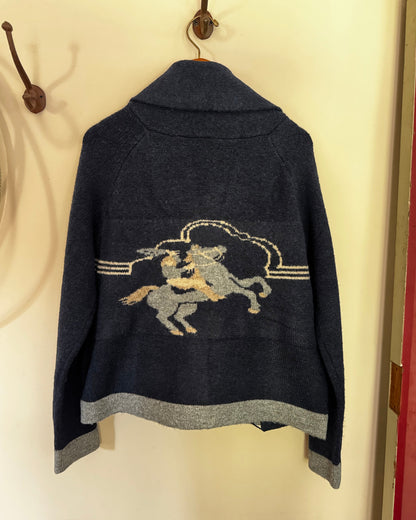 A dark navy blue knit sweater with a gray and beige embroidered cowboy riding a rearing horse design on the back, hanging on a hook against a cream-colored wall.