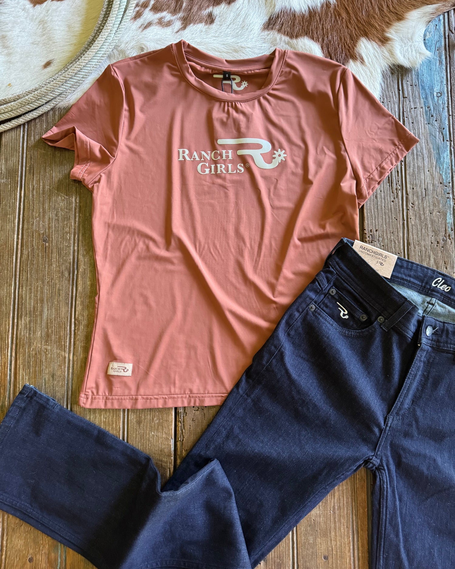 Pink t-shirt with 'Ranch Girls' logo and dark blue jeans on a wooden surface.
