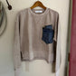 Beige sweatshirt with a blue pocket on a wooden hanger against a white wall.