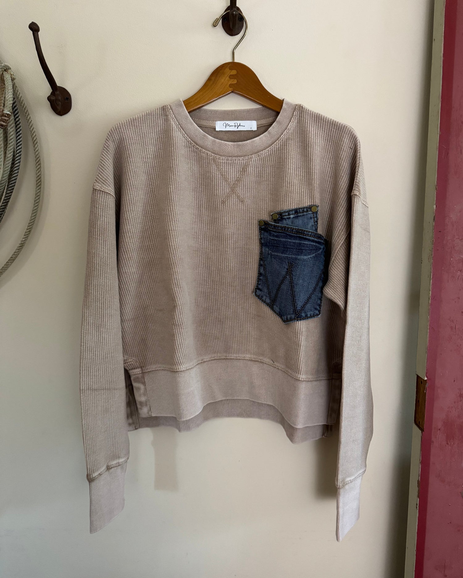 Beige sweatshirt with a blue pocket on a wooden hanger against a white wall.