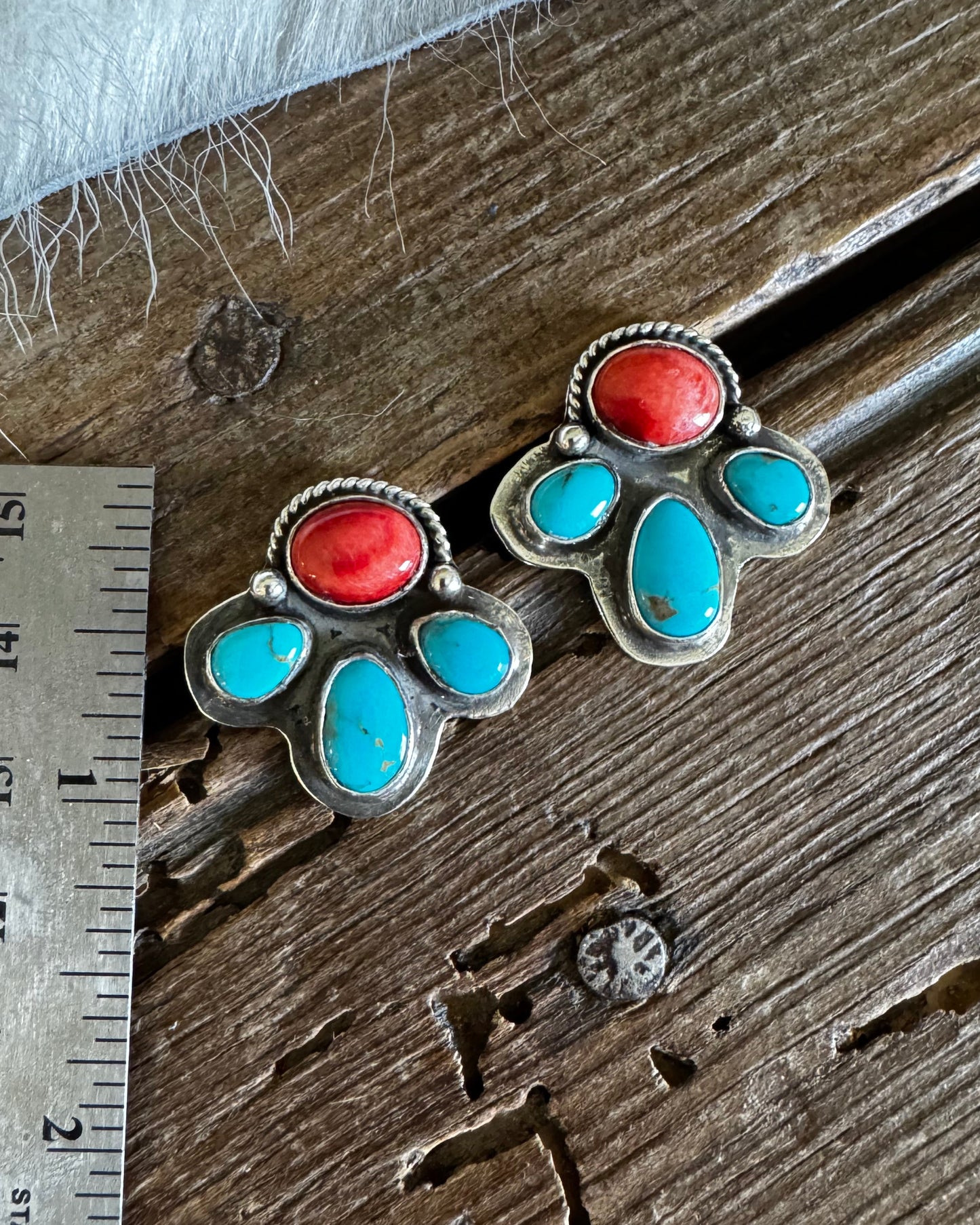 A pair of silver-toned paw-shaped earrings with turquoise and coral stone inlays, displayed on a rustic wooden surface next to a metal ruler for scale.