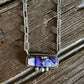 A silver-toned chain necklace with a rectangular bezel pendant containing purple and white marbled stone, displayed on a rustic dark wood surface.