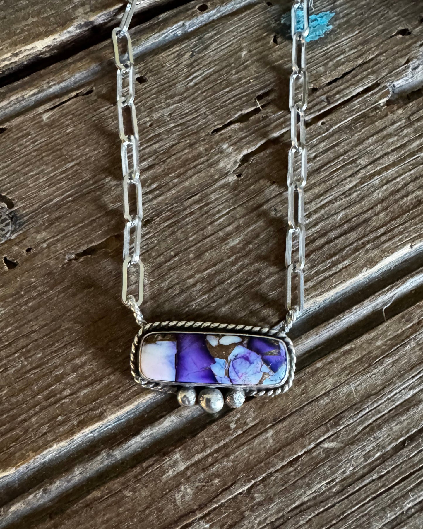 A silver-toned chain necklace with a rectangular bezel pendant containing purple and white marbled stone, displayed on a rustic dark wood surface.