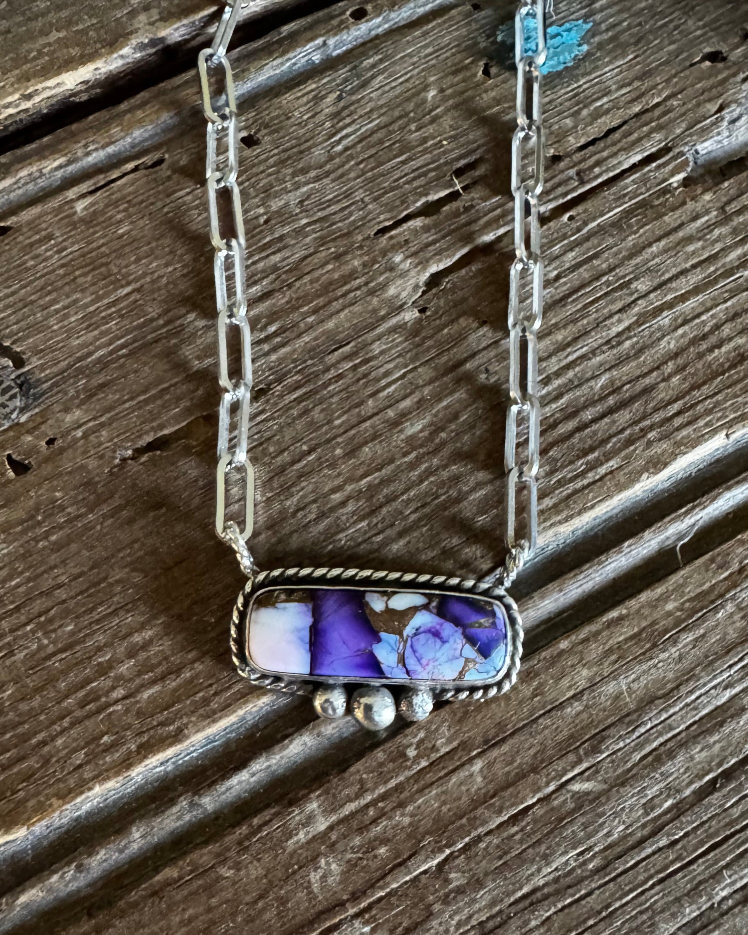 A silver-toned chain necklace with a rectangular bezel pendant containing purple and white marbled stone, displayed on a rustic dark wood surface.