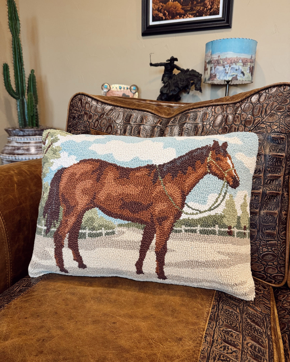 A rectangular woven throw pillow featuring a brown horse illustration, resting on a brown leather and embossed leather armchair in a living room setting.