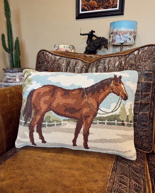 A rectangular woven throw pillow featuring a brown horse illustration, resting on a brown leather and embossed leather armchair in a living room setting.