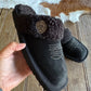 Black fuzzy slippers with a brand logo held by a hand on a wooden floor.