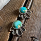 A pair of silver-toned statement earrings with oval turquoise-blue stones and a scalloped petal-like metal border, displayed on a rustic wooden surface with a white fur trim in the corner.