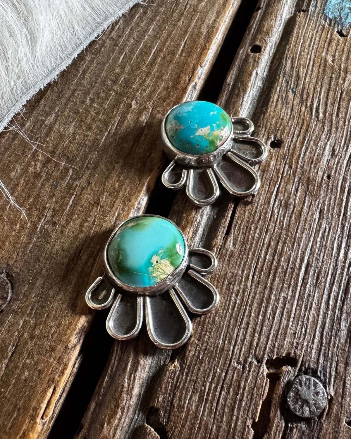 A pair of silver-toned statement earrings with oval turquoise-blue stones and a scalloped petal-like metal border, displayed on a rustic wooden surface with a white fur trim in the corner.