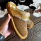 A pair of tan suede slip-on slippers with a fluffy cream shearling-style strap and embroidered floral detail, held in a hand over a cowhide rug and dark wood floor.