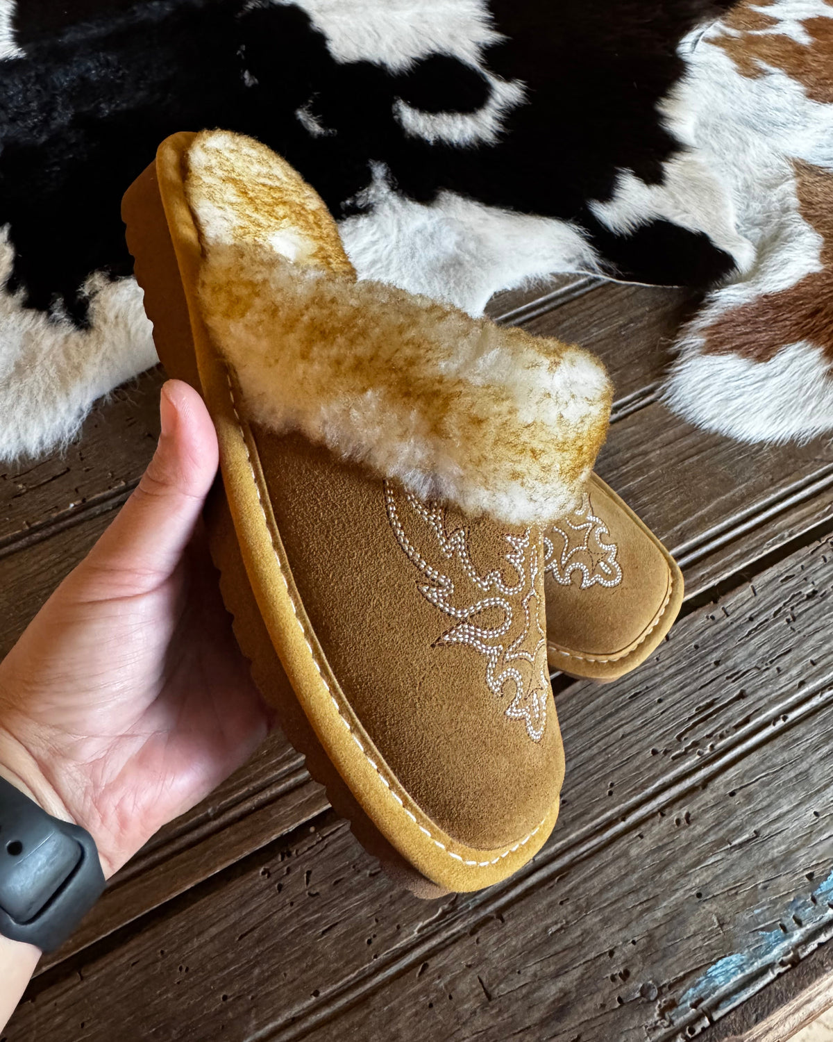 A pair of tan suede slip-on slippers with a fluffy cream shearling-style strap and embroidered floral detail, held in a hand over a cowhide rug and dark wood floor.