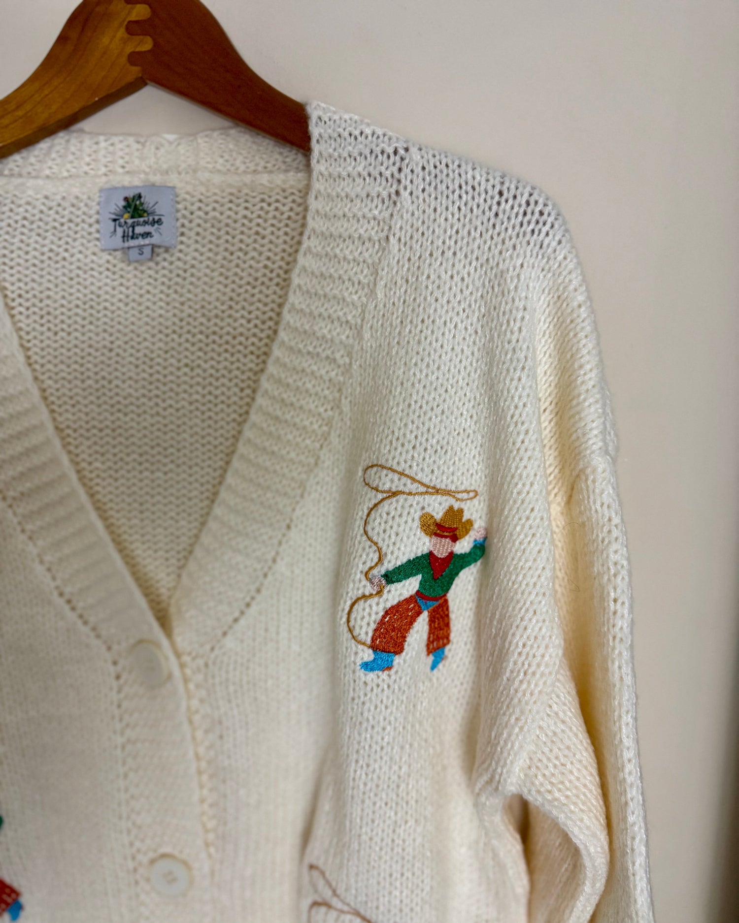 White cardigan with embroidered designs on a wooden hanger against a white background