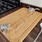 A rectangular bamboo cutting board with a carved handle and two silver metal bull skull handles, resting on a dark wood kitchen countertop next to a stove.