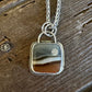 A silver-toned necklace with a square pendant featuring a natural agate or stone pattern in earthy brown, gray, and cream tones, displayed on a rustic dark wood surface.