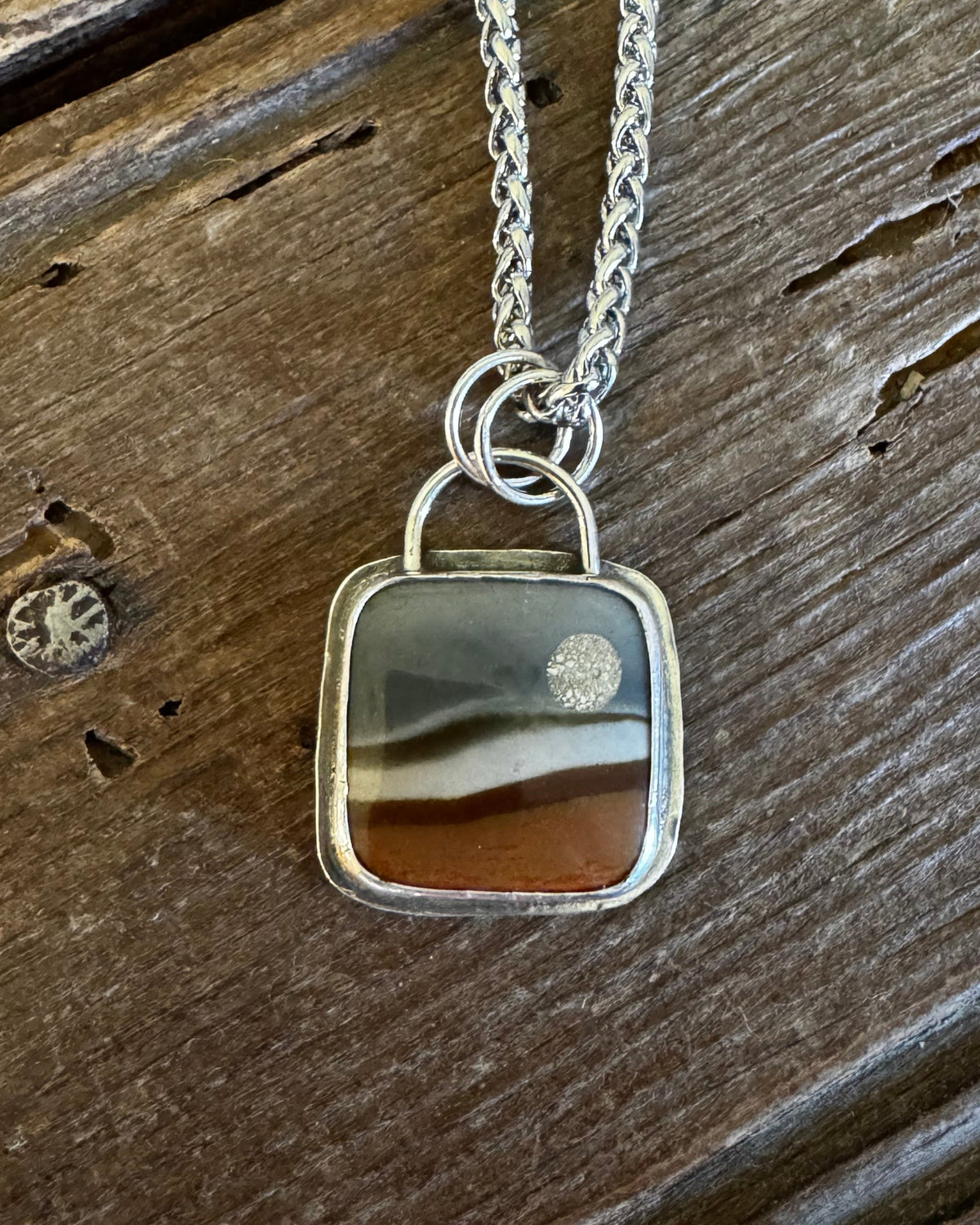 A silver-toned necklace with a square pendant featuring a natural agate or stone pattern in earthy brown, gray, and cream tones, displayed on a rustic dark wood surface.