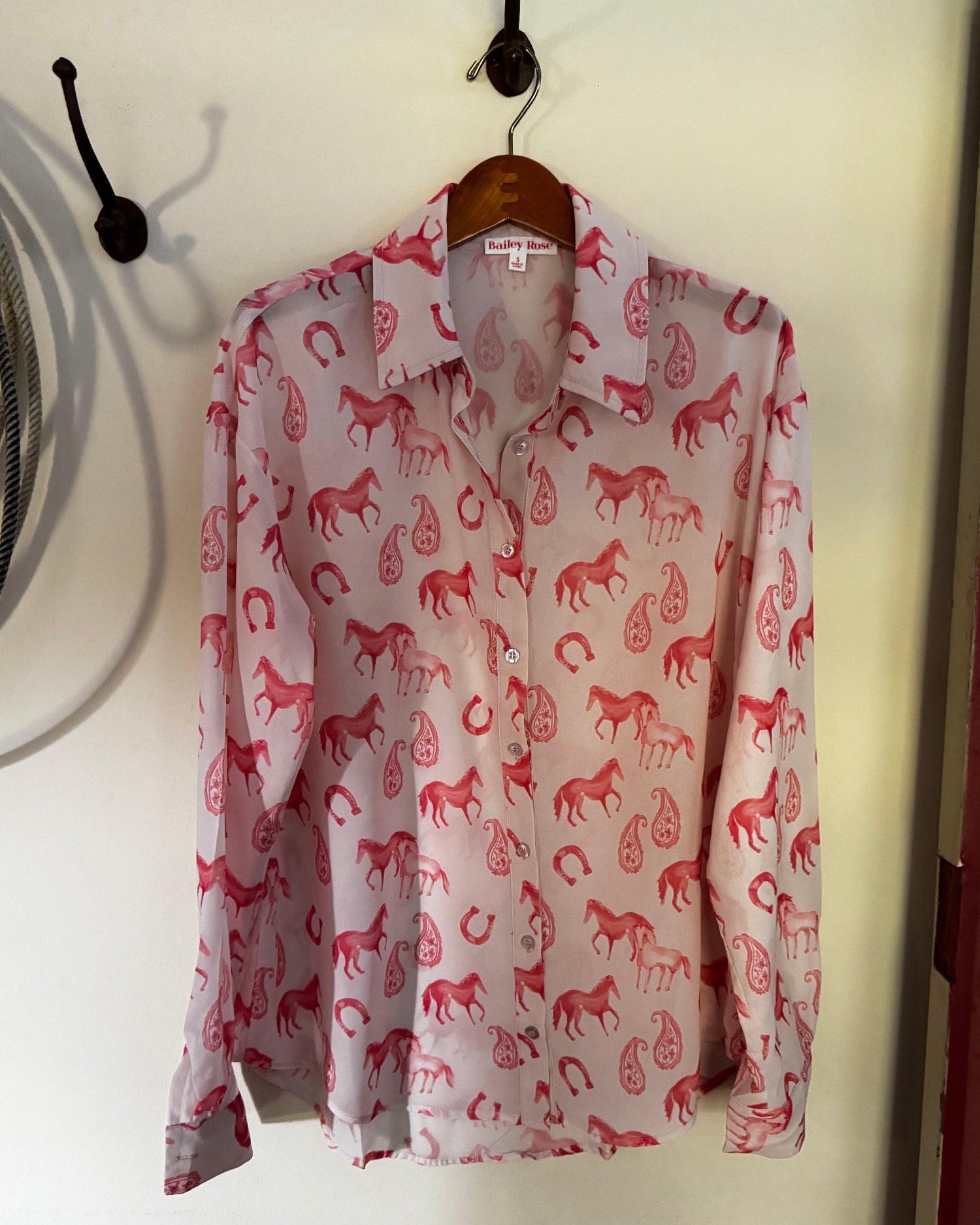 A long-sleeve button-up shirt with an allover red horse and rider pattern, hanging on a wooden hanger against a white wall.