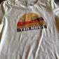 White t-shirt with a graphic design featuring a sunset and cactus, labeled 'Tin Haul', on a wooden surface.