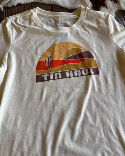White t-shirt with a graphic design featuring a sunset and cactus, labeled 'Tin Haul', on a wooden surface.