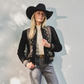 Woman wearing a black embroidered jacket, beige sweater, and jeans with a black hat against a light gray background.
