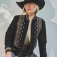 Person wearing a black embroidered jacket and cowboy hat sitting on a stool against a light background