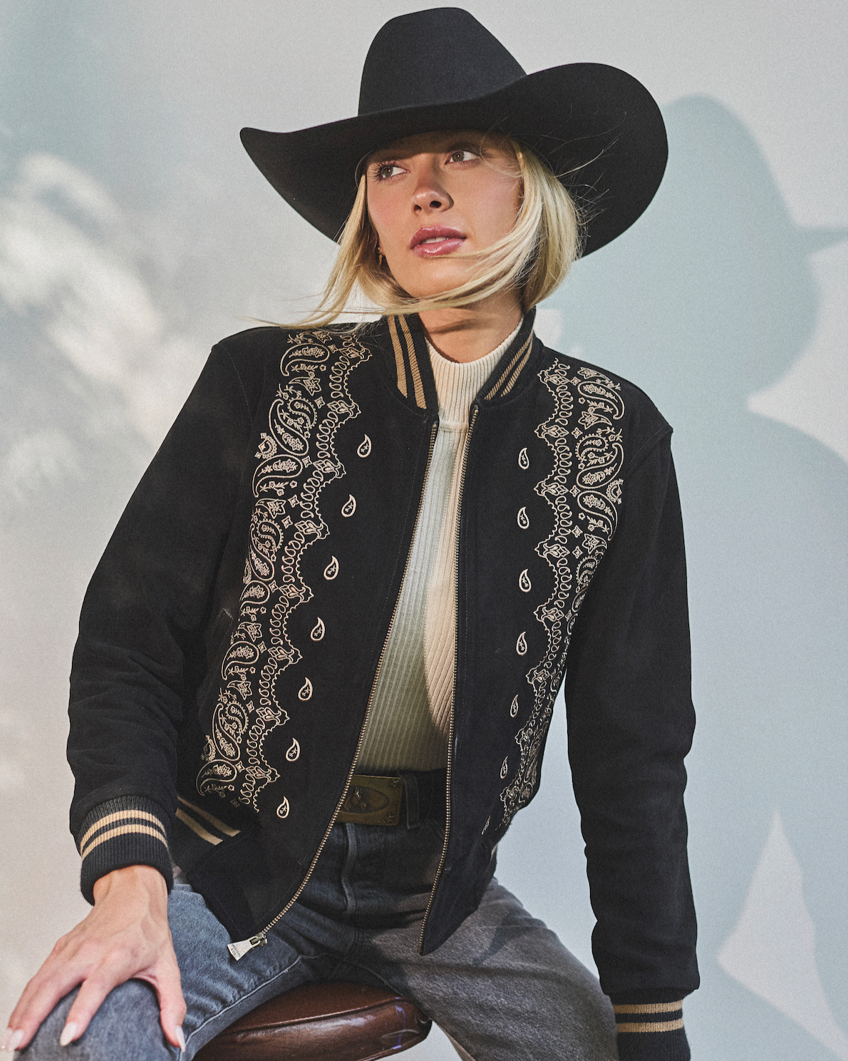 Person wearing a black embroidered jacket and cowboy hat sitting on a stool against a light background