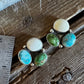 A pair of handmade sterling silver stud earrings with mixed natural stones (turquoise, green, cream, and brown) displayed on a rustic wooden surface next to a metal ruler for scale.