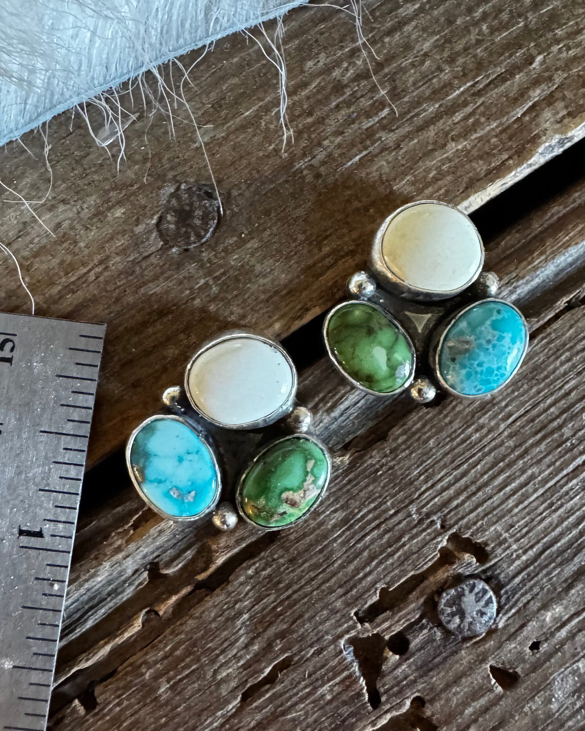 A pair of handmade sterling silver stud earrings with mixed natural stones (turquoise, green, cream, and brown) displayed on a rustic wooden surface next to a metal ruler for scale.