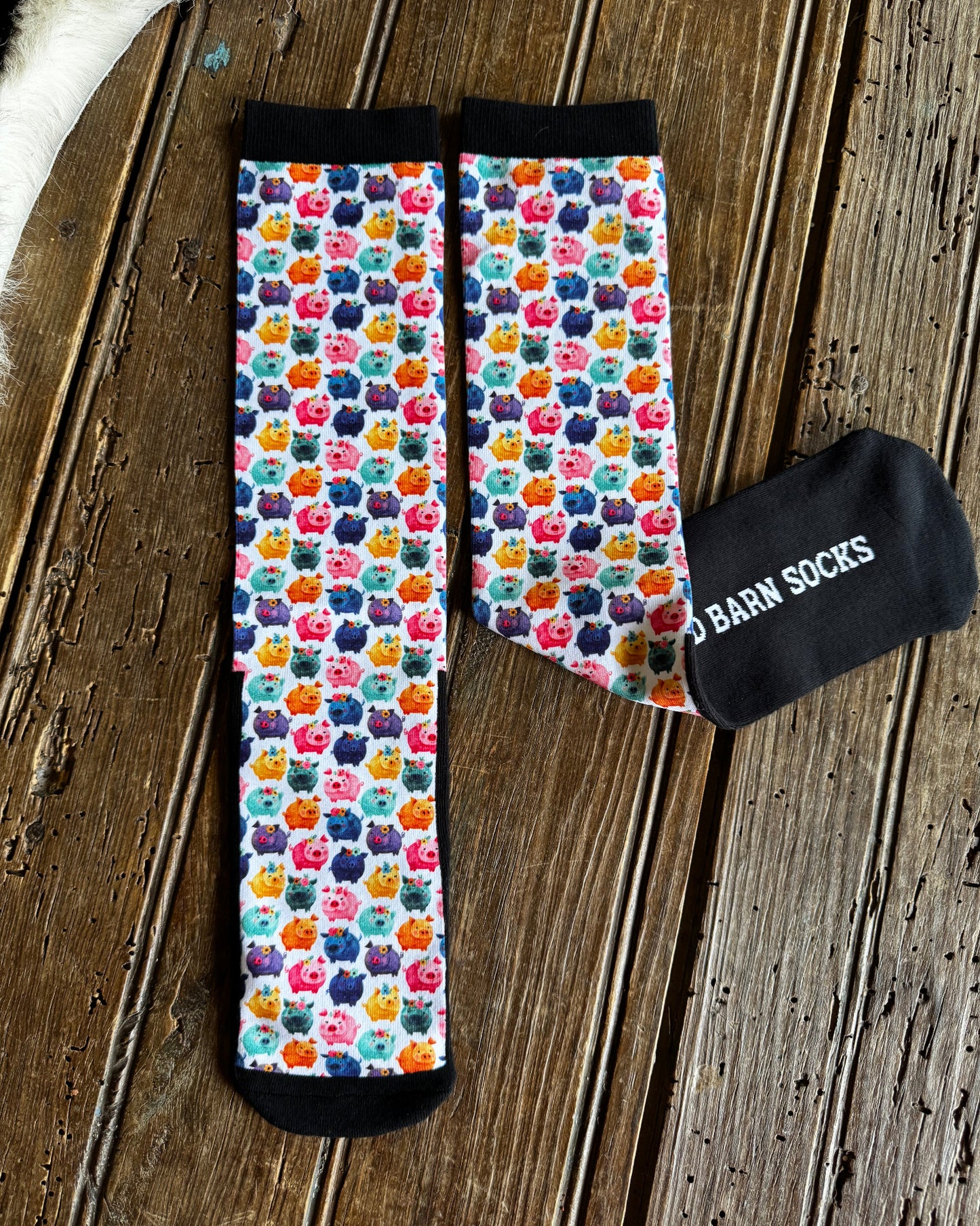 A pair of crew socks with a colorful repeating pattern of cartoon pigs on a white background, finished with solid black cuffs and toes. One sock is laid flat showing the pattern, while the other is folded to show the black sole with white text reading 'BARN SOCKS'.