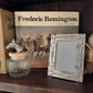 Decorative items on a wooden shelf including a book titled 'Frederic Remington', a picture frame, and a glass jar.