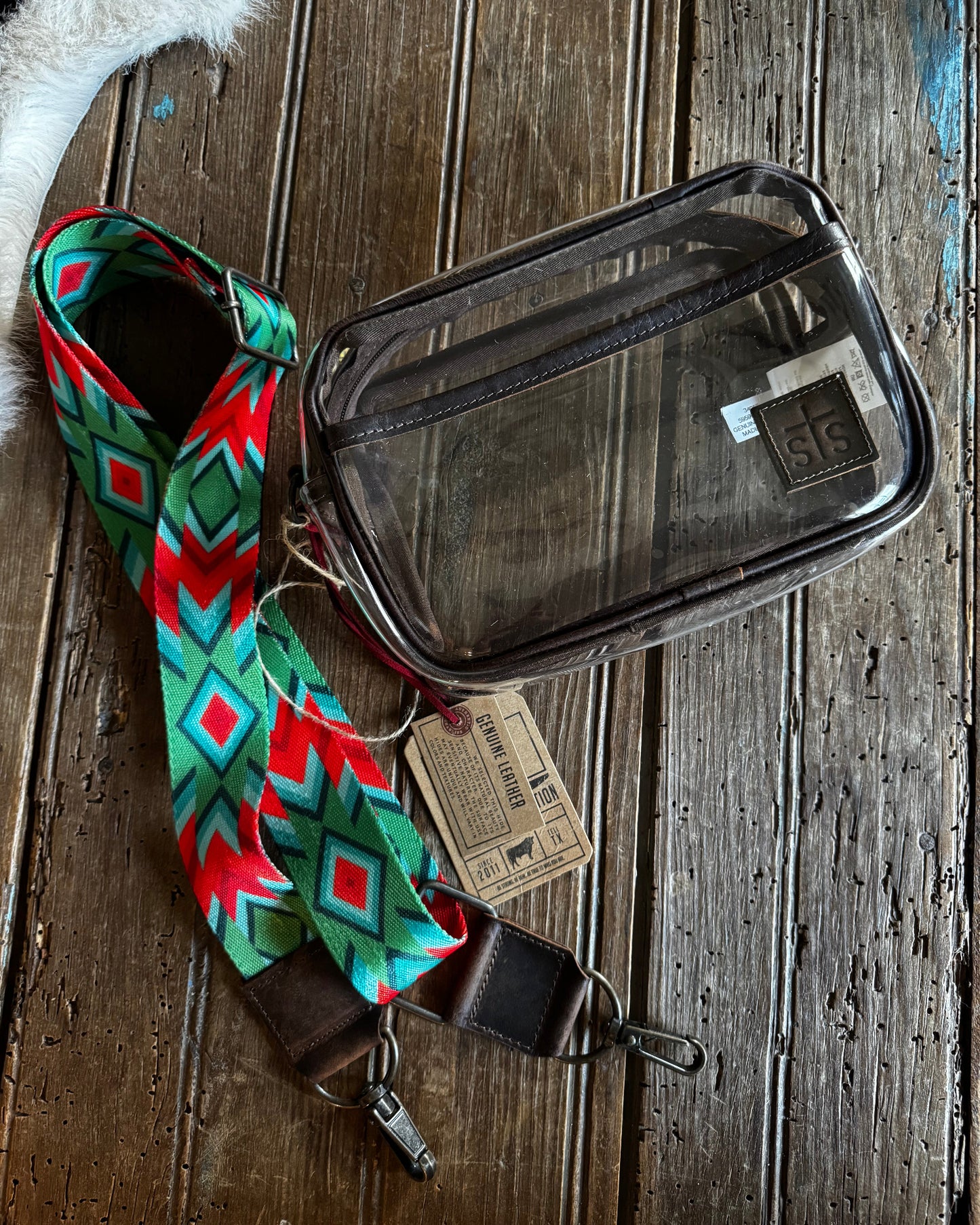 A clear plastic zippered pouch with a brown leather trim and a colorful geometric patterned webbing strap, displayed on a rustic wooden surface.