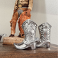 Silver cowboy boot figurines on a wooden surface with a statue in the background.