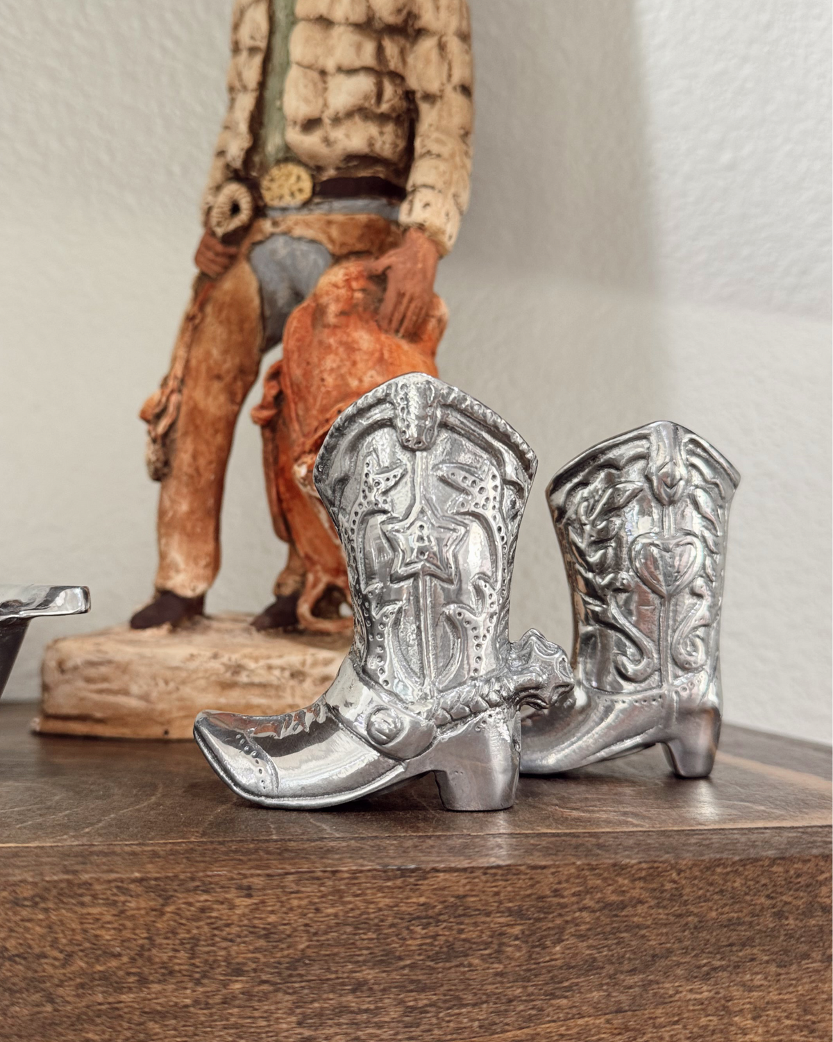 Silver cowboy boot figurines on a wooden surface with a statue in the background.