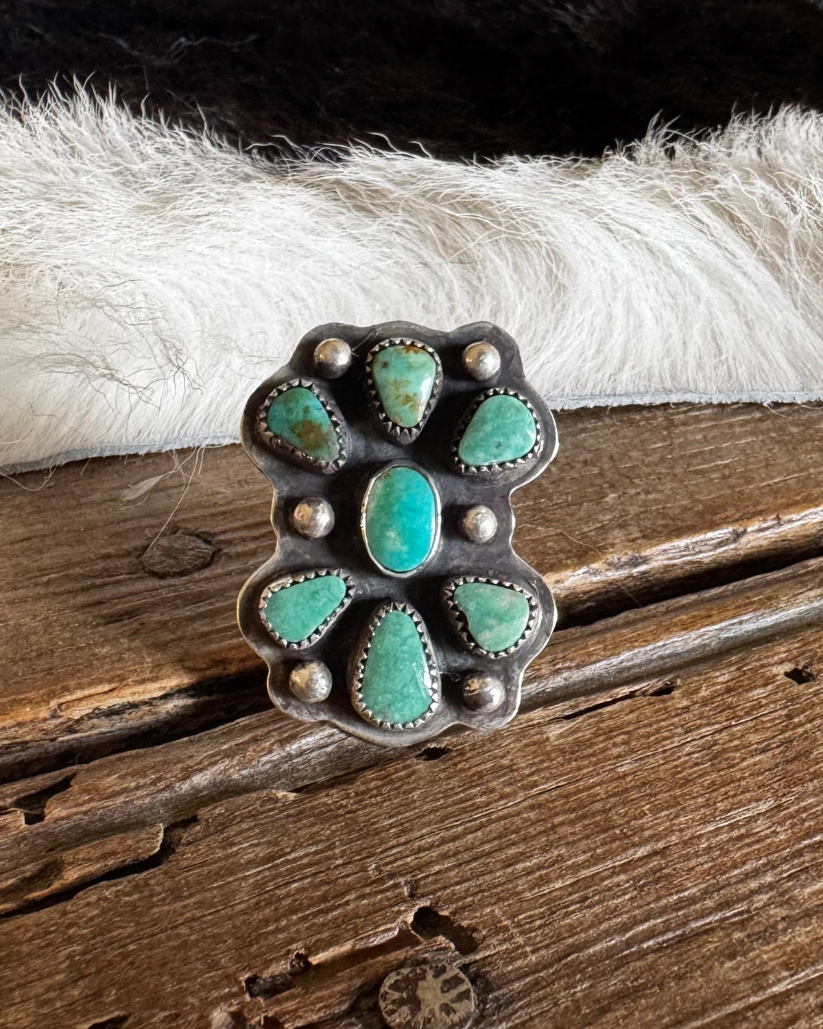 A sterling silver ring with a floral cluster of turquoise stones, displayed on a rustic wooden surface with a black and white cowhide rug behind it.