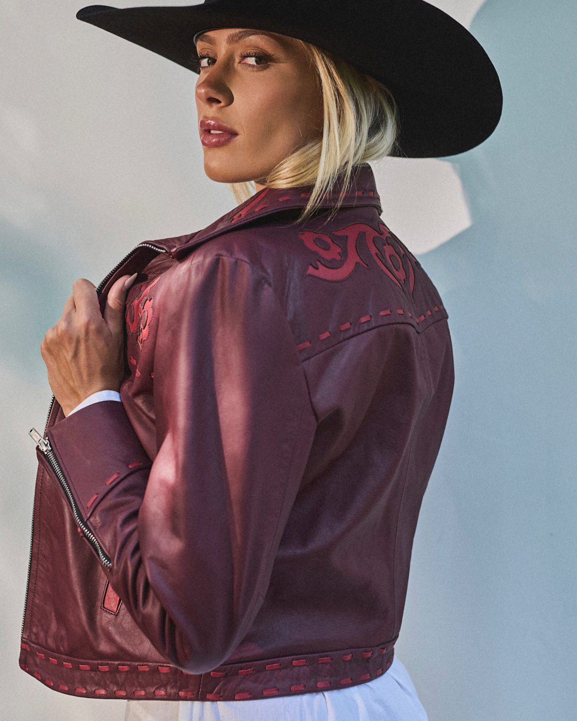 Woman wearing a burgundy leather jacket with red embroidery and a black cowboy hat against a light gray background
