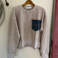 Beige sweatshirt with a blue pocket on a hanger against a white wall.